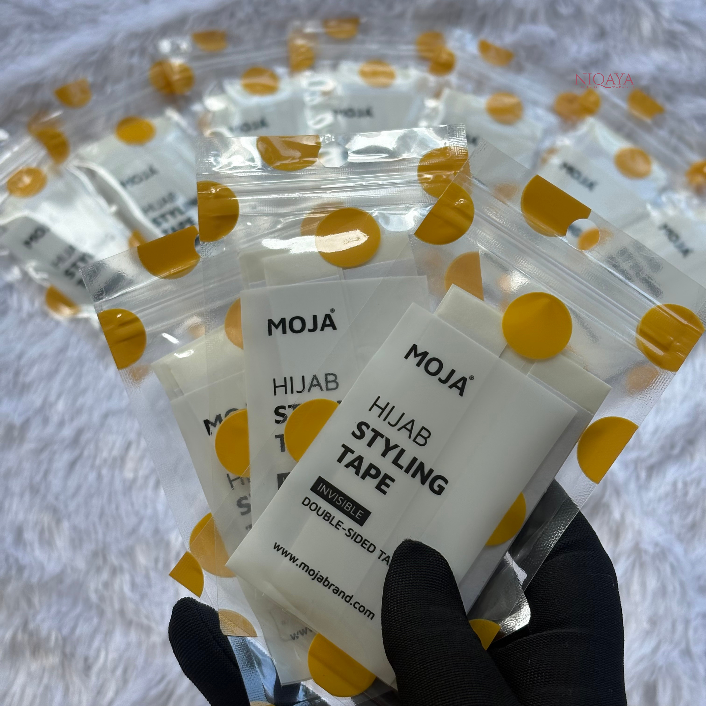MOJA Hijab Styling Tape packages held by a hand with black gloves on a white background