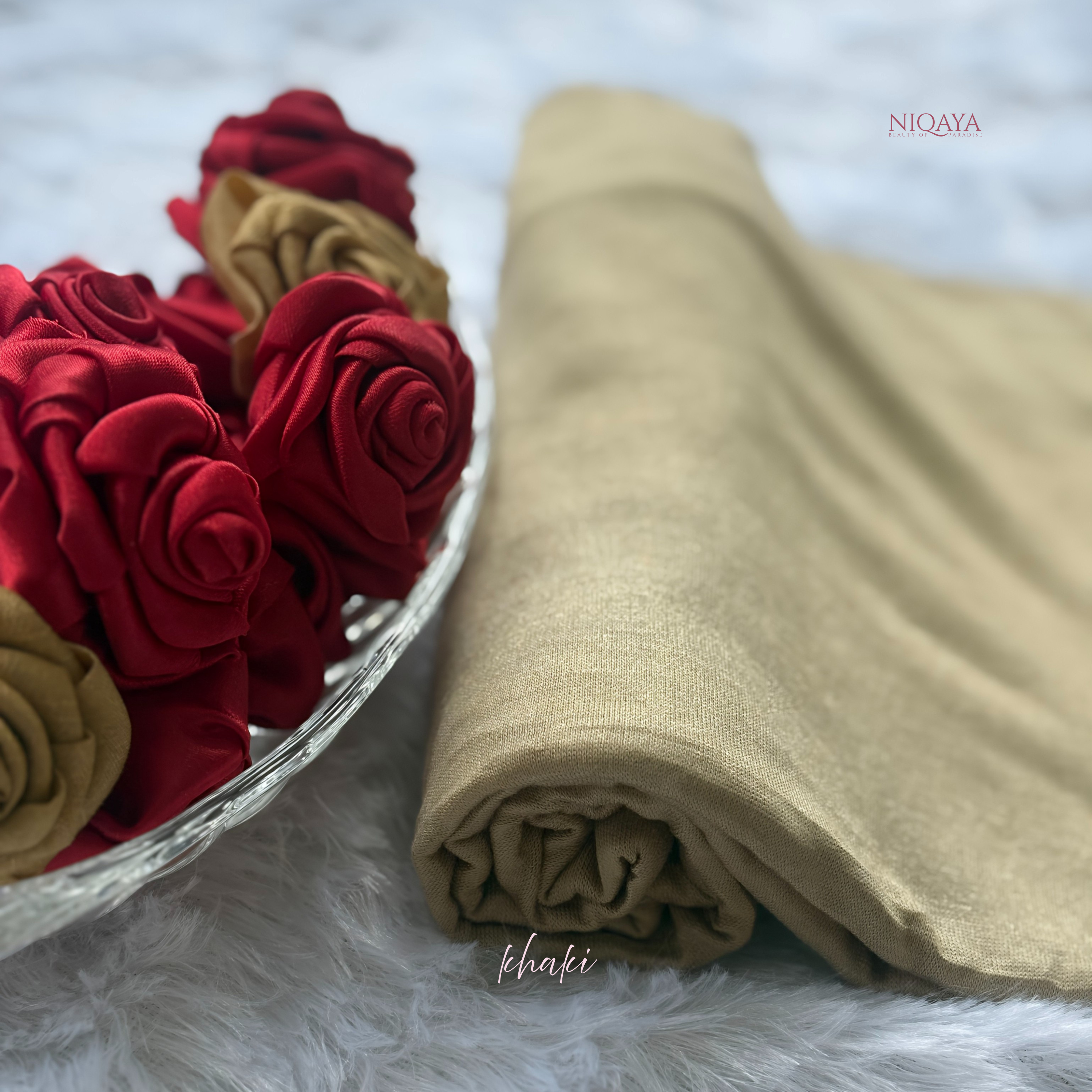 Folded khaki fabric next to red and gold fabric flowers on a white surface with 'NIQAYA' branding.