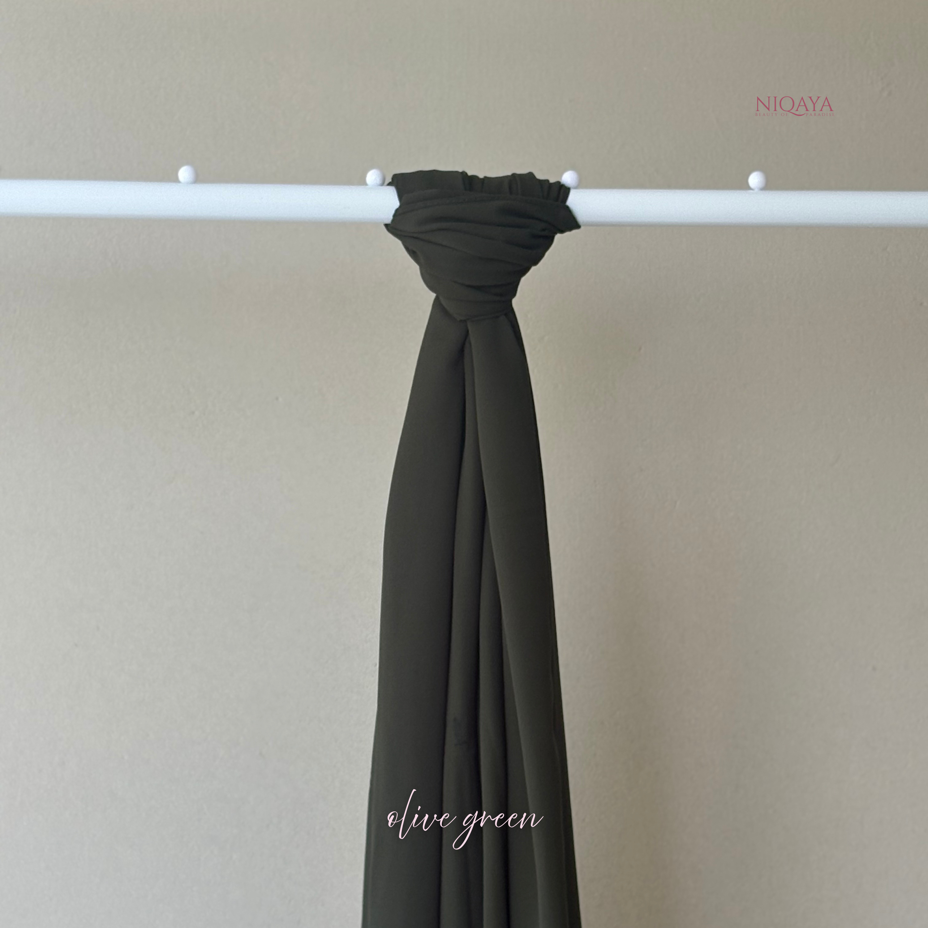 Olive green scarf draped over a white rod against a beige background, with 'Niqaya' branding.