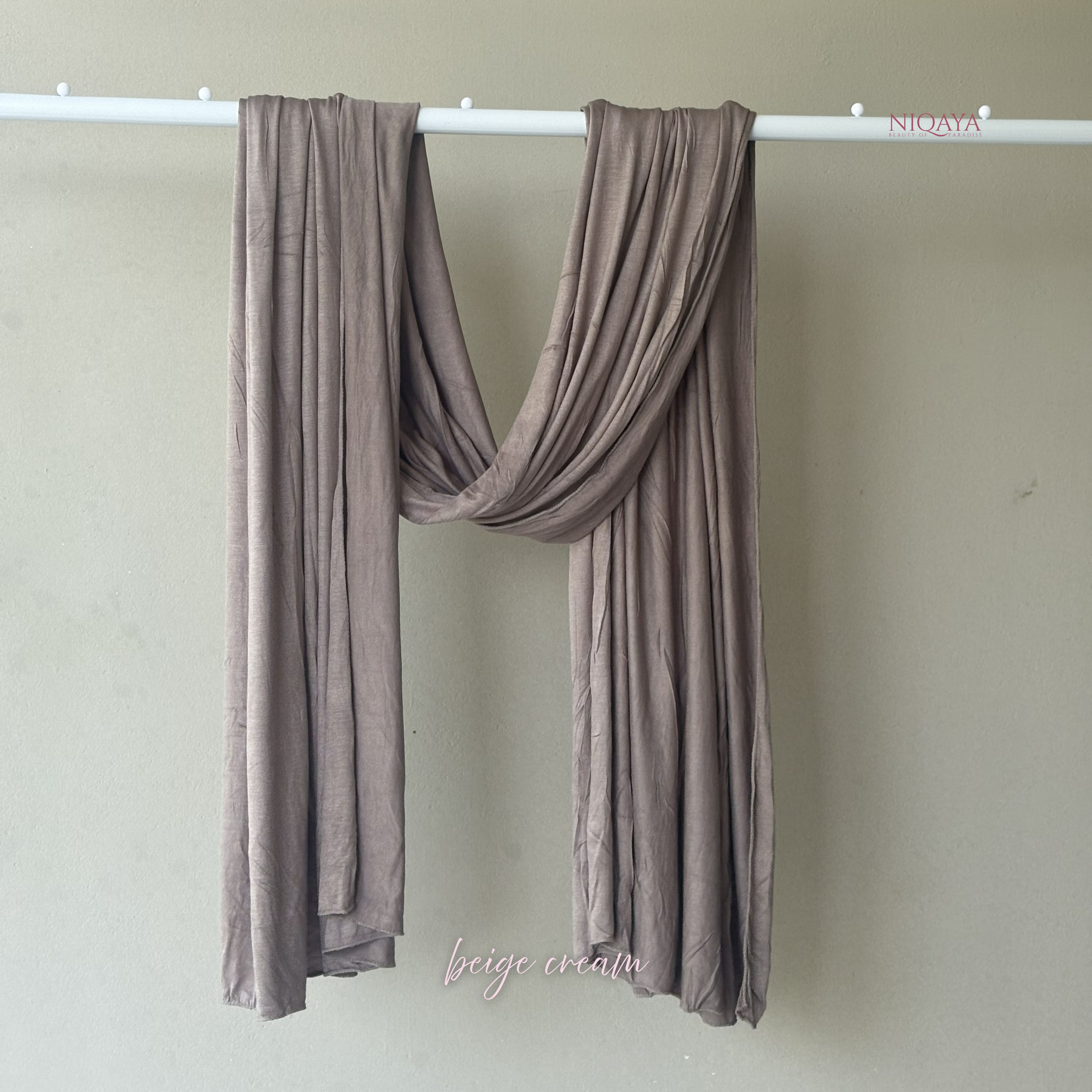 Brown scarf draped over a white rod against a beige wall with 'Niqaa' branding.