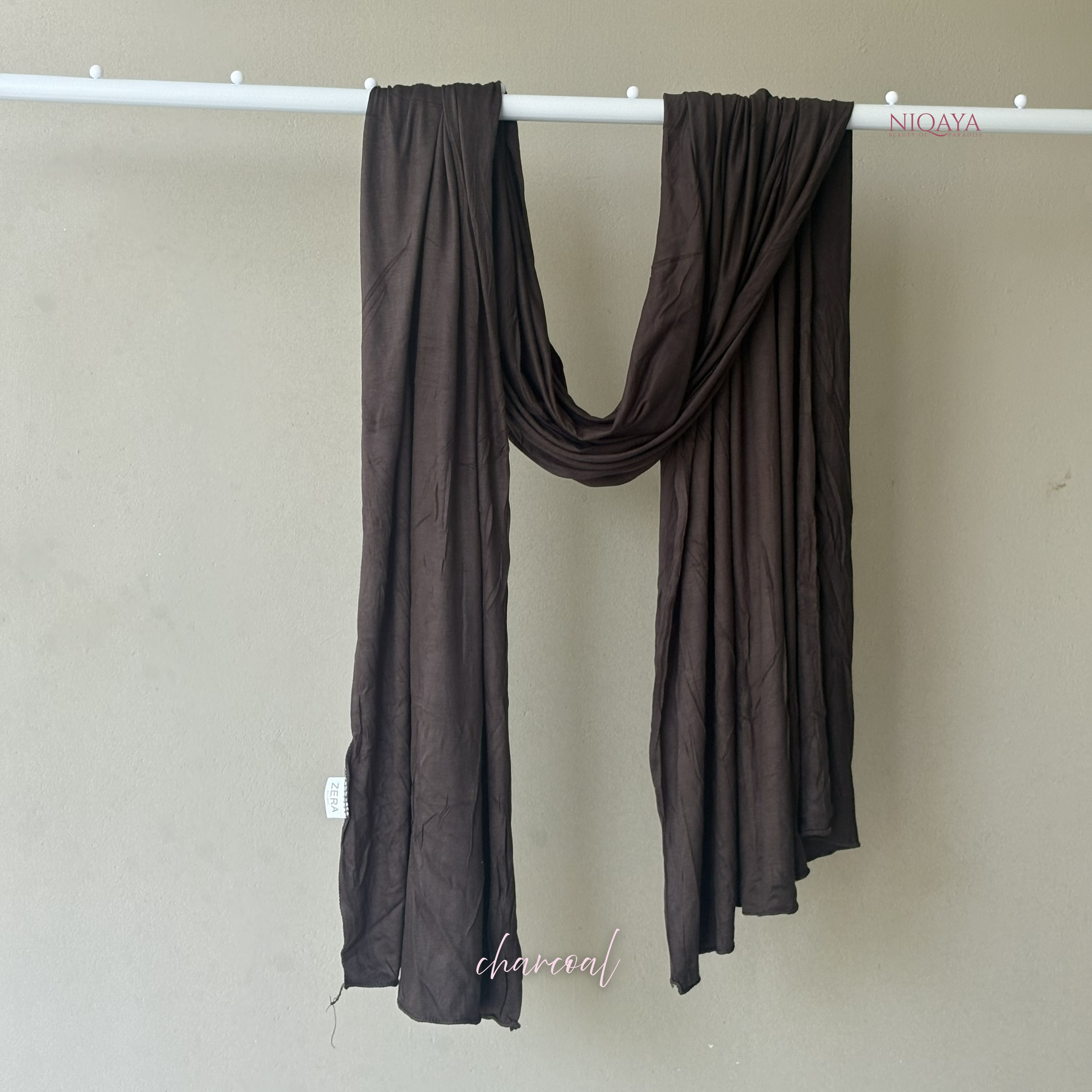 Brown scarf draped over a white rod against a beige wall with 'NIOQA' branding.