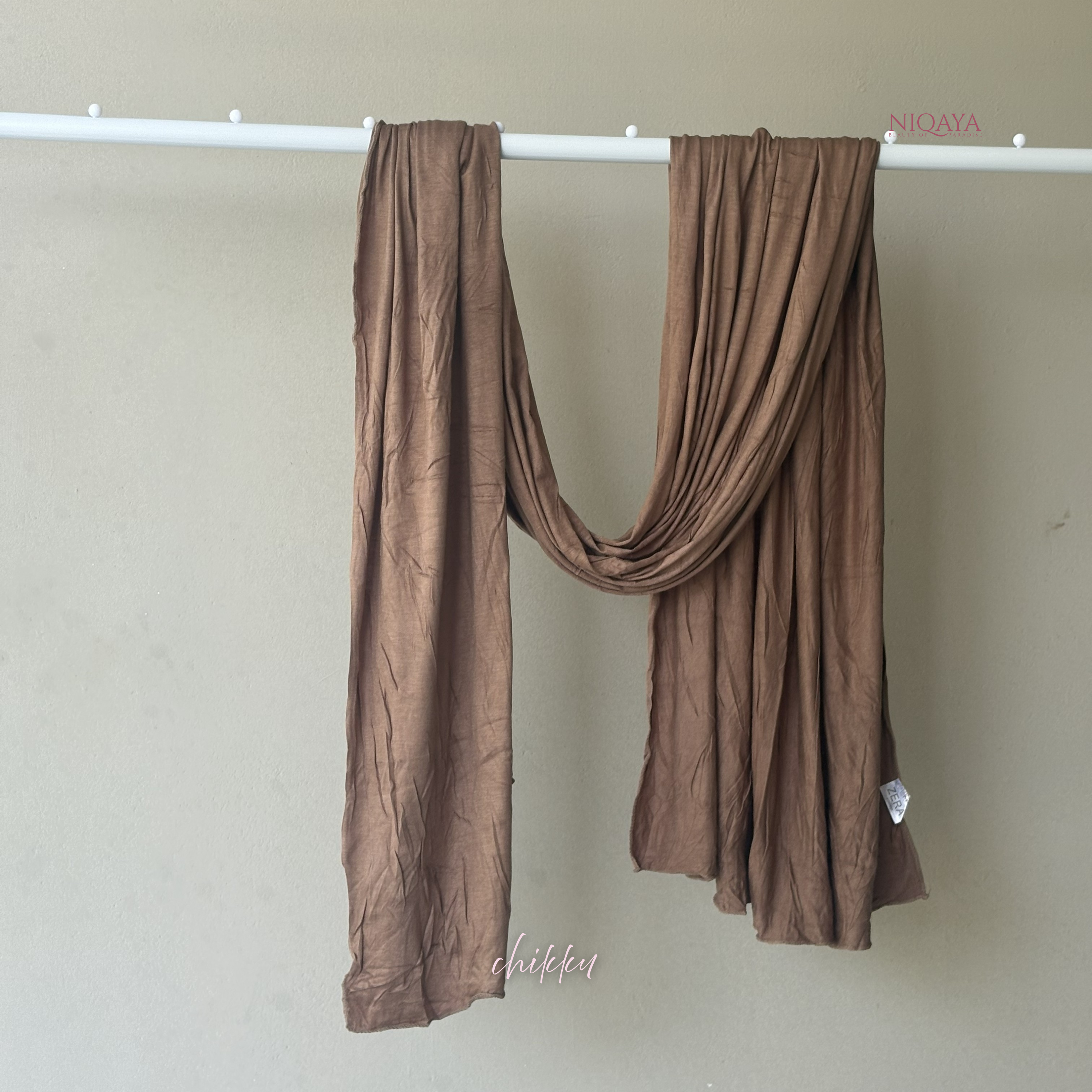 Brown scarf draped over a white rod against a beige wall with 'Niqaya' branding.