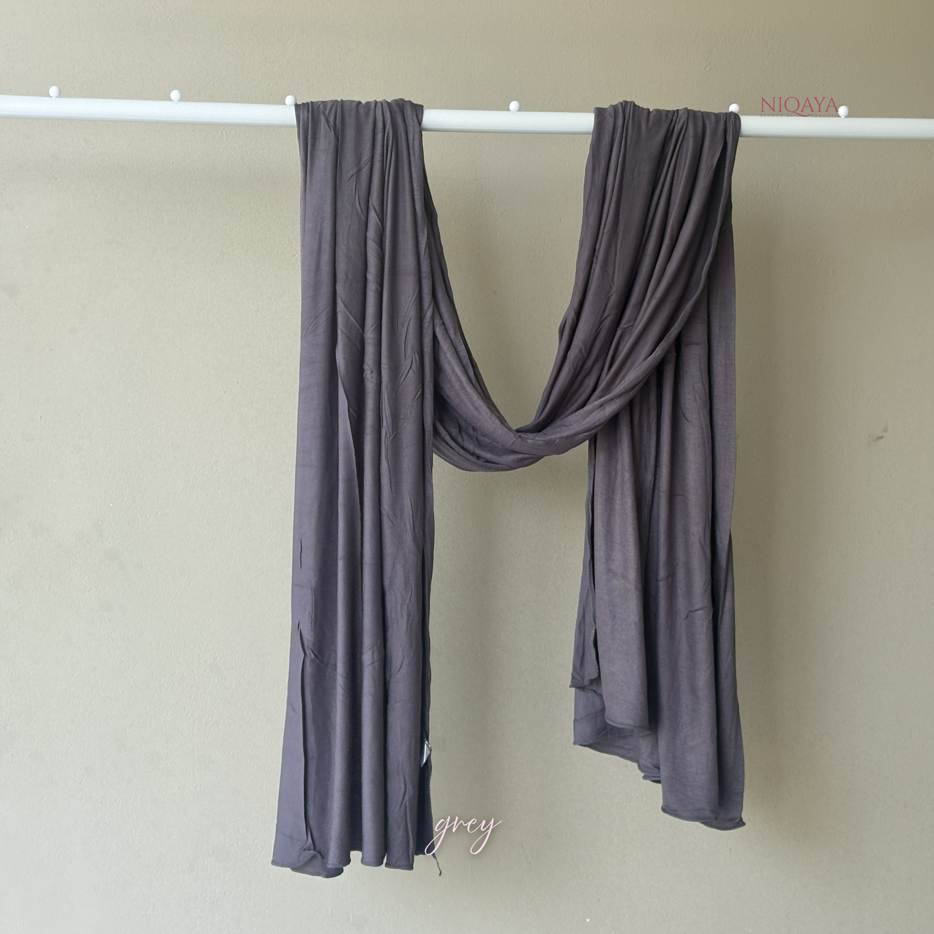 Gray scarf draped over a white rod against a beige wall, with 'Nioaya' branding.