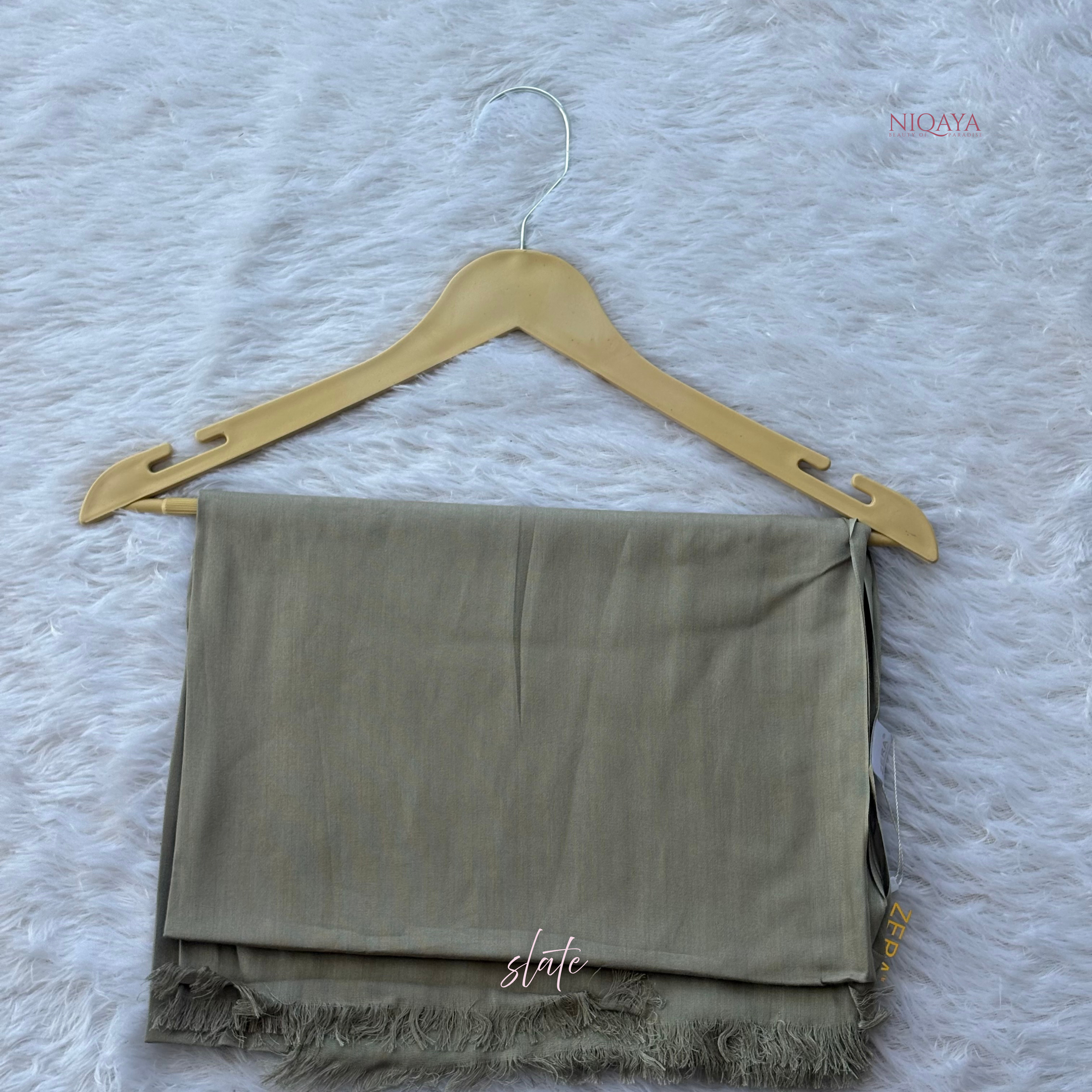 Folded green scarf on a wooden hanger against a textured gray background, with 'Niqaya' branding.